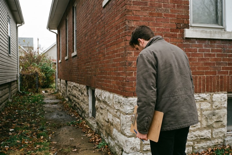 An engineer with a clipboard looking at the foundation of a brick bungalow